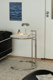 A modern round side table with a clear glass top and a curved chrome metal frame, styled in a living room setting with a silver dome lamp and a book on top.