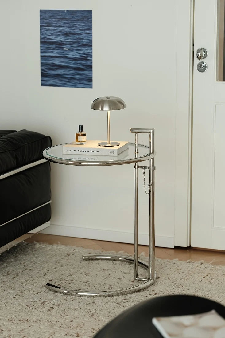 A modern round side table with a clear glass top and a curved chrome metal frame, styled in a living room setting with a silver dome lamp and a book on top.