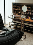 A modern round glass side table with a chrome metal frame and adjustable lamp, styled in a contemporary living room next to a black leather sofa and TV stand.