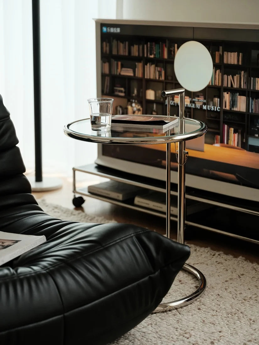 A modern round glass side table with a chrome metal frame and adjustable lamp, styled in a contemporary living room next to a black leather sofa and TV stand.