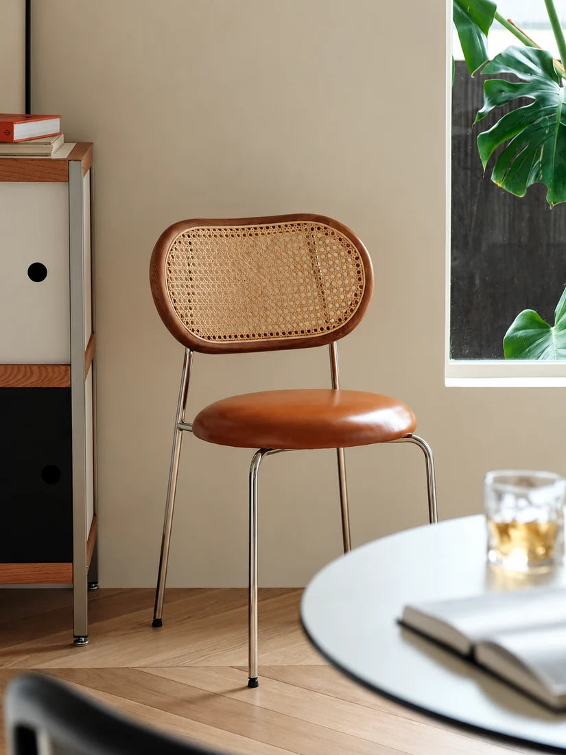 A mid-century modern dining chair with a curved brown leather seat and a woven cane backrest, set in a warm, minimalist room next to a window with green leaves.