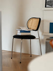 A modern black chair with a woven cane backrest and chrome legs, styled with a stack of books on the seat in a minimalist room with light wood flooring.