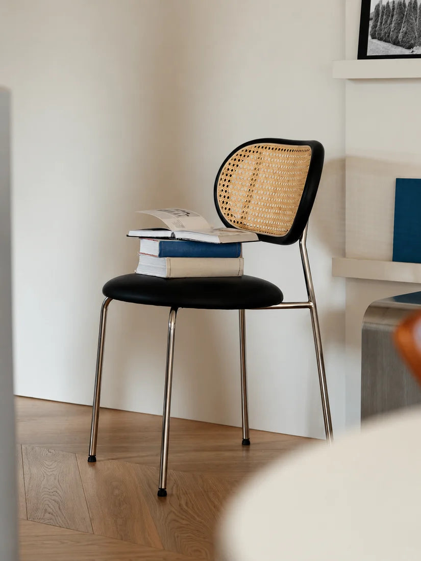 A modern black chair with a woven cane backrest and chrome legs, styled with a stack of books on the seat in a minimalist room with light wood flooring.