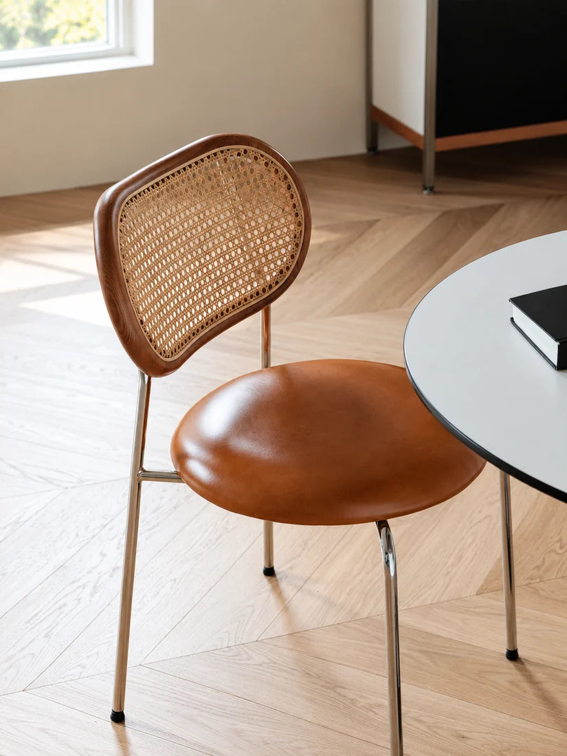 A mid-century modern dining chair with a curved brown leather seat and backrest, featuring a woven cane back panel and slim metal legs, shown in a bright room with a light wood herringbone floor and a white table edge.