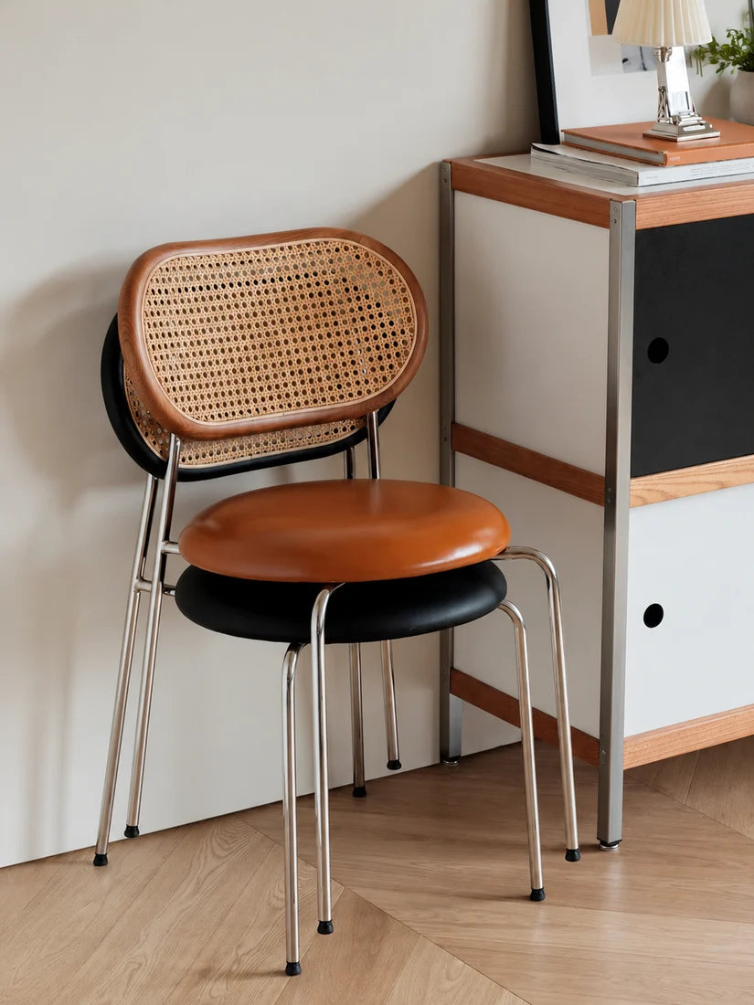 A modern stackable chair with a woven cane backrest, brown leather seat, and black cushion, set in a minimalist room next to a wooden cabinet.