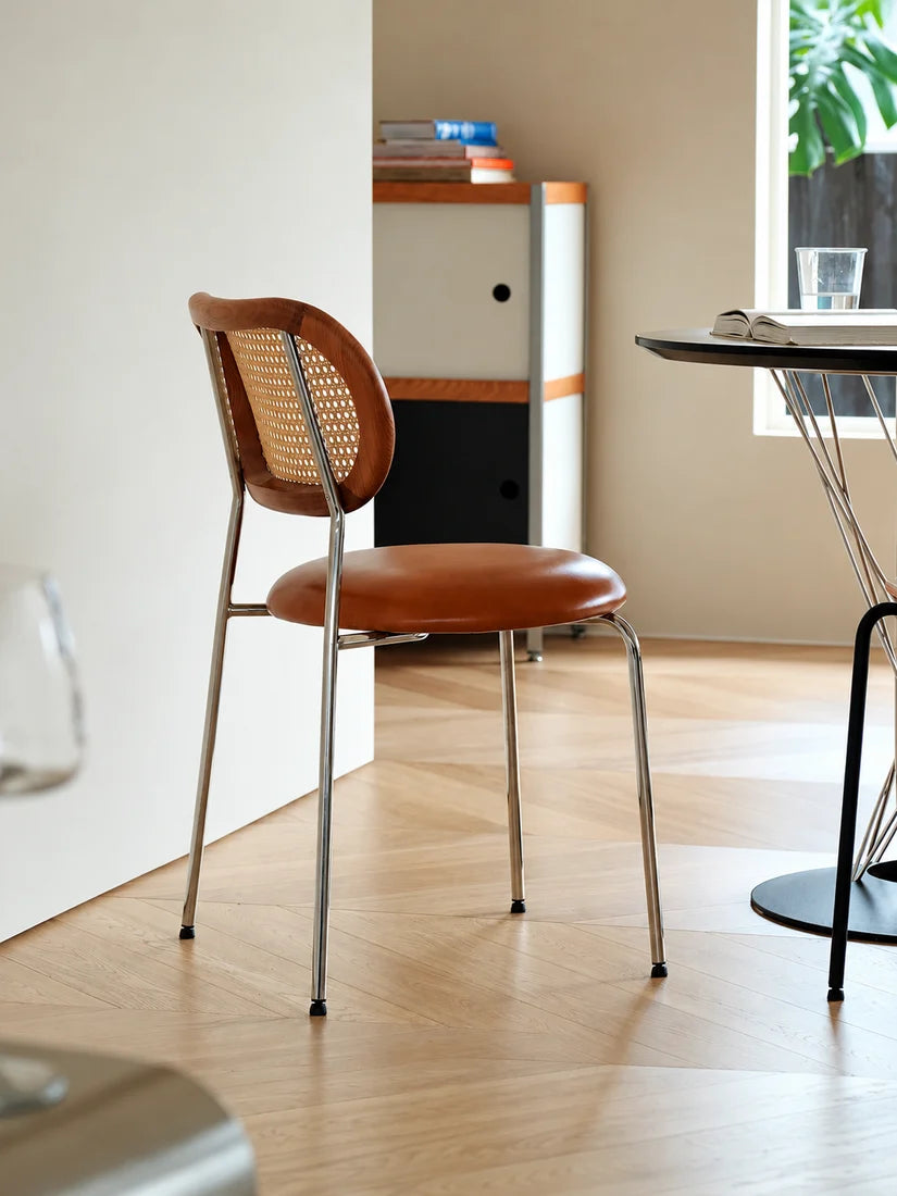 A mid-century modern dining chair with a brown leather seat and a woven cane backrest, set in a bright, contemporary room with light wood floors.