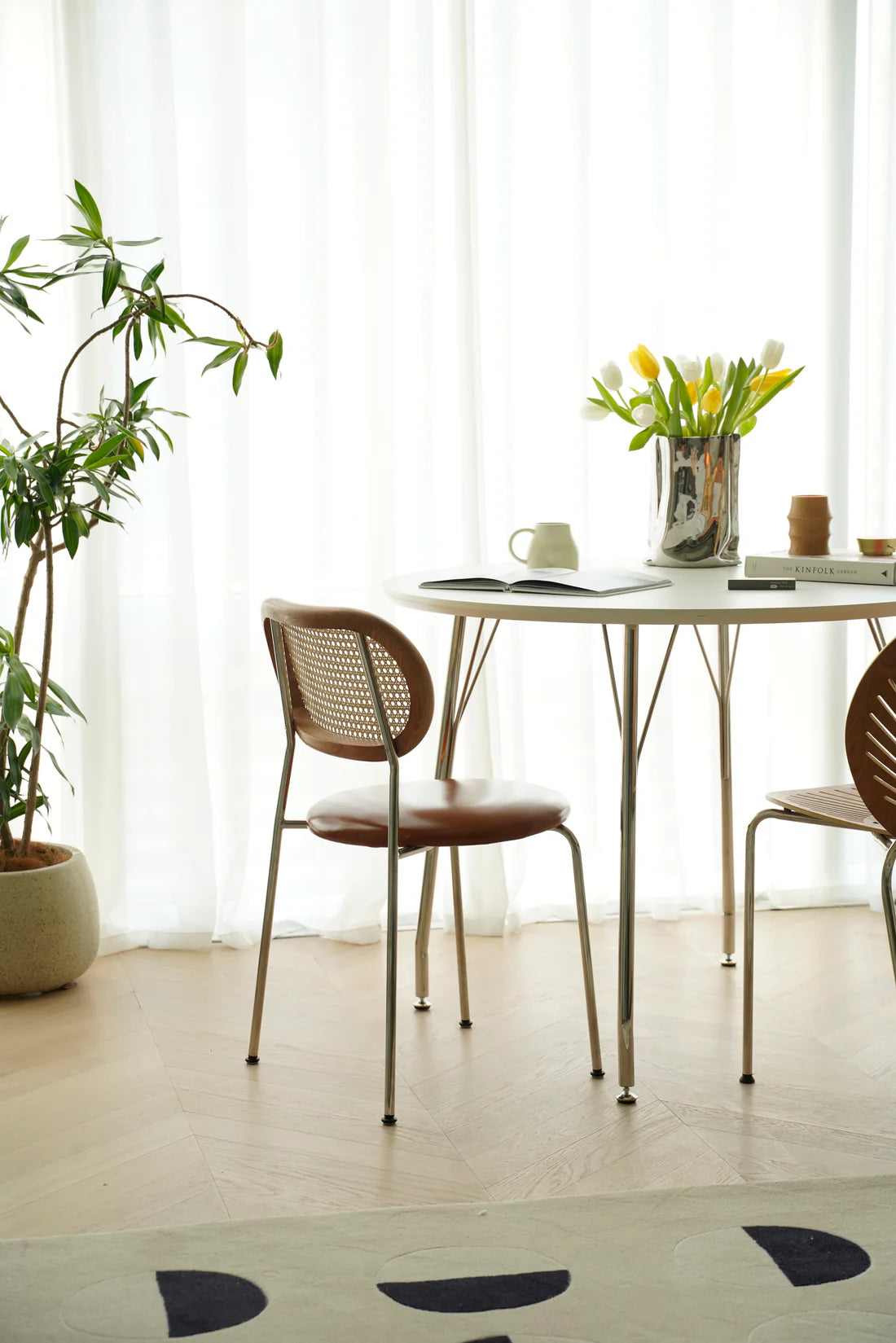 Modern dining area with a small table and wooden rattan chairs near a window with white curtains.