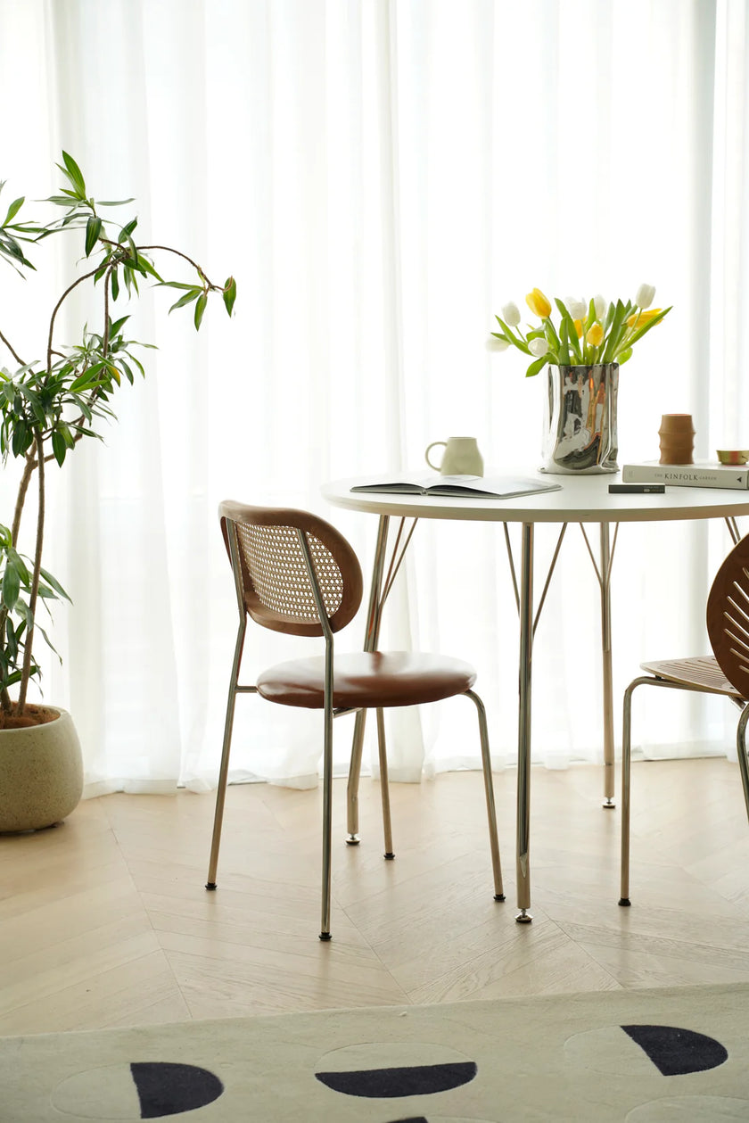 Modern dining area with a small table and wooden rattan chairs near a window with white curtains.