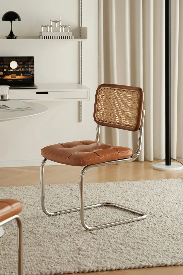 A modern side chair with a brown leather seat and backrest, featuring a woven cane back panel and a chrome cantilever base, set in a bright, contemporary room on a textured cream rug.