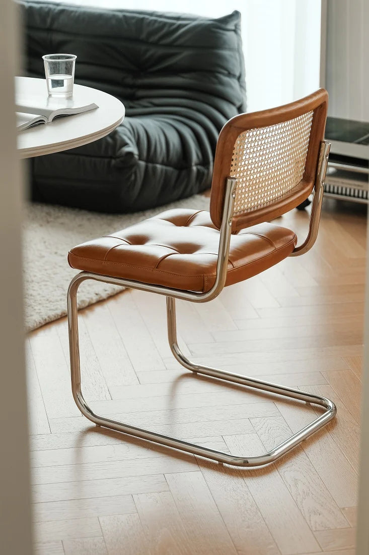 A modern cantilever dining chair with a tufted tan leather seat and backrest, featuring a woven cane back panel and a polished chrome metal base, shown in a bright living room setting.