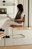 A woman sits on a modern sling chair with a brown leather seat and woven back, reading a book in a bright, minimalist living room.