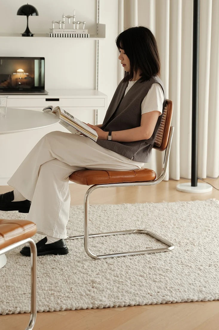 A woman sits on a modern sling chair with a brown leather seat and woven back, reading a book in a bright, minimalist living room.