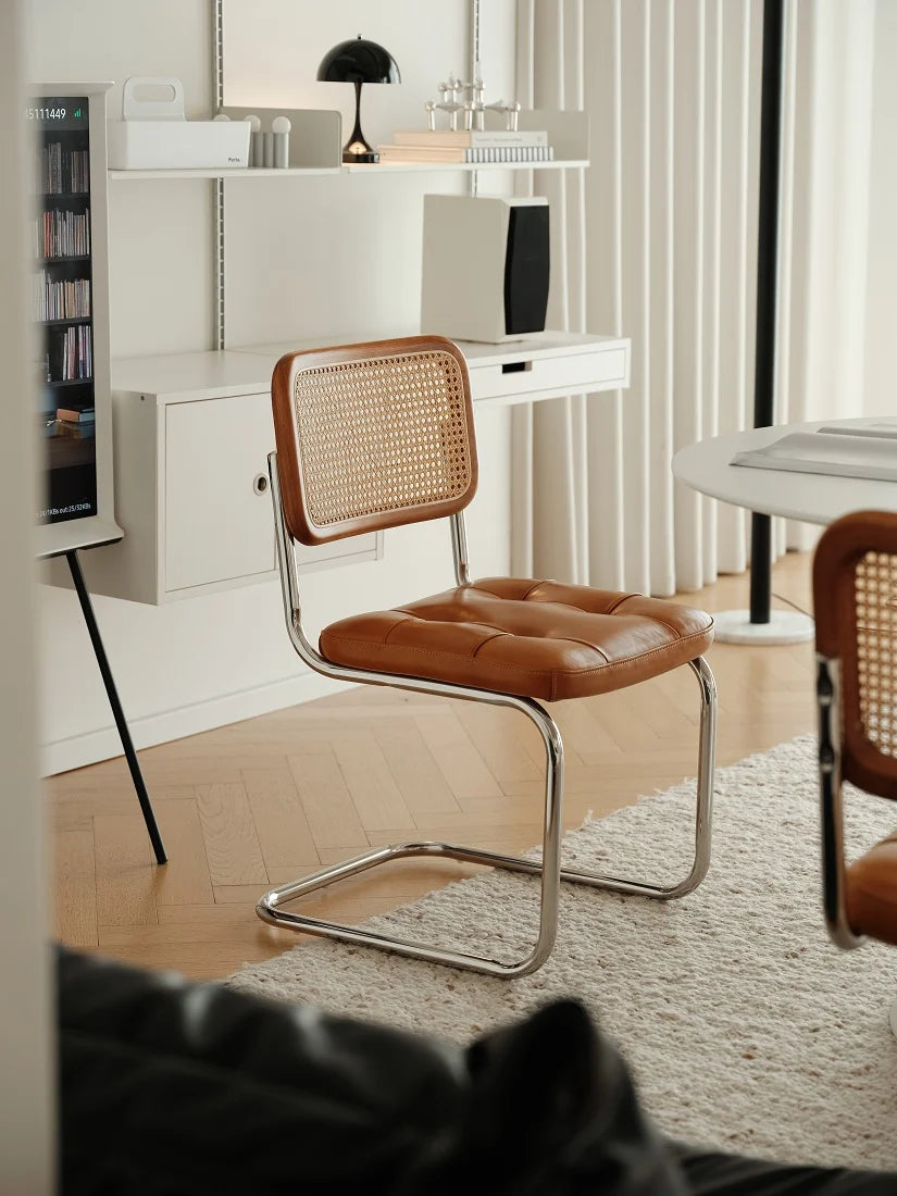 A modern cantilever chair with a brown leather seat and a woven cane backrest, positioned in a bright, contemporary room with light wood floors and white furniture.
