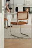 A modern cantilever chair with a brown leather seat and a woven cane backrest, positioned on a textured light gray rug in a bright, contemporary room.
