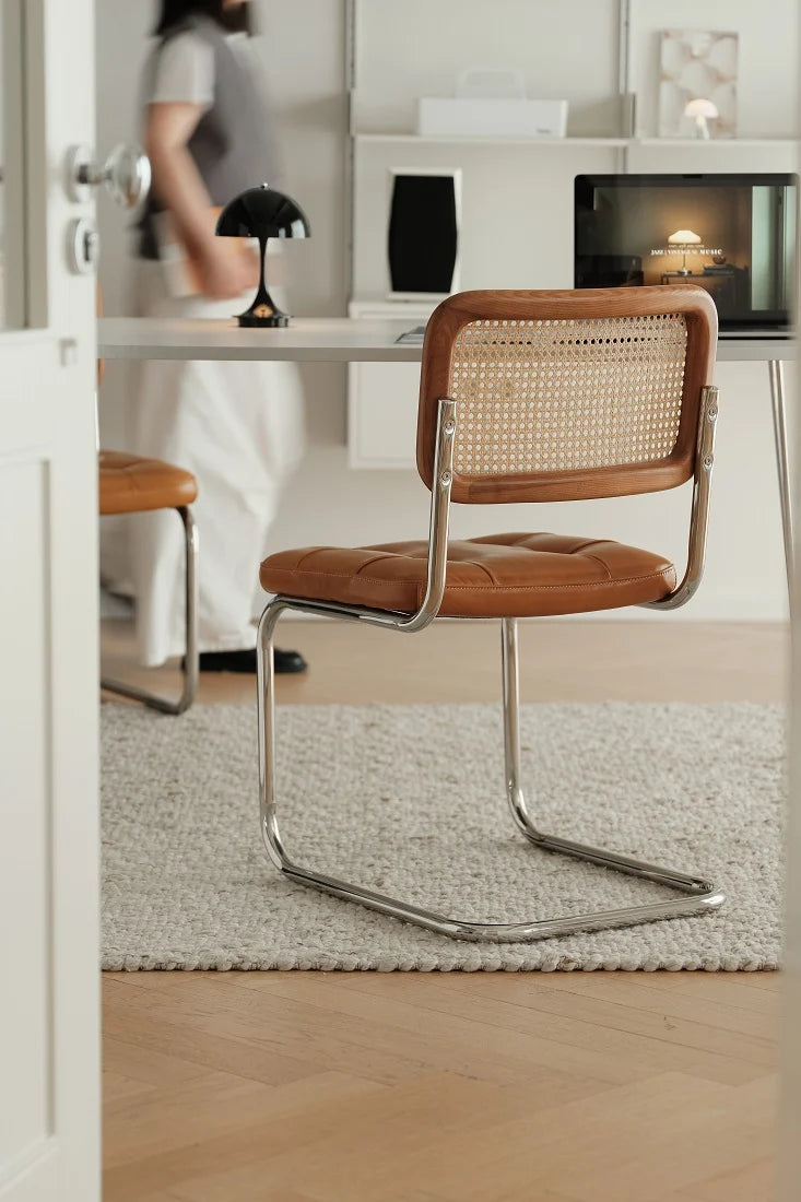 A modern cantilever chair with a brown leather seat and a woven cane backrest, positioned on a textured light gray rug in a bright, contemporary room.