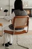 A woman sits on a modern chair with a brown leather seat and backrest featuring a woven cane back panel, reading a book in a bright, contemporary room.