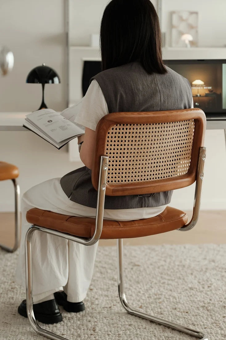 A woman sits on a modern chair with a brown leather seat and backrest featuring a woven cane back panel, reading a book in a bright, contemporary room.