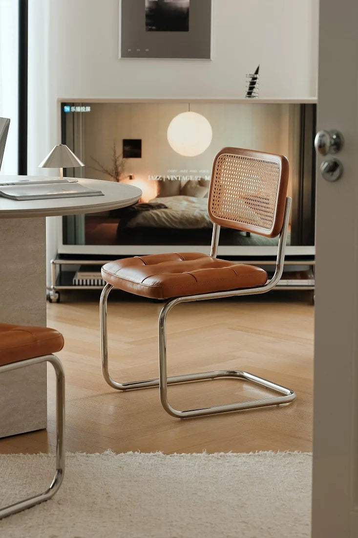 A modern cantilever chair with a brown leather seat and backrest woven cane back, shown in a stylishly decorated room with a wooden floor and a large mirror.