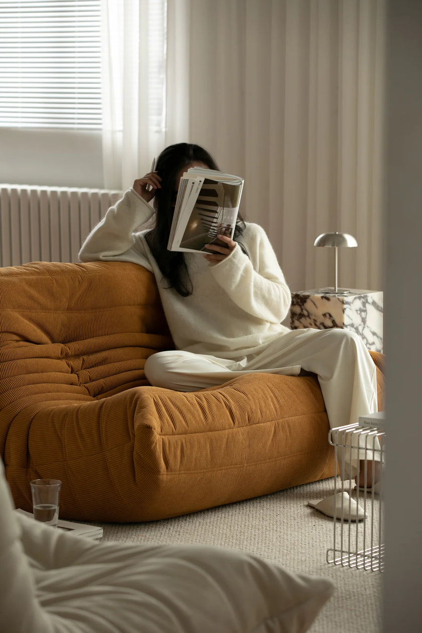 Women sitting on a mustard yellow corduroy sofa reading a book in a cozy living room.