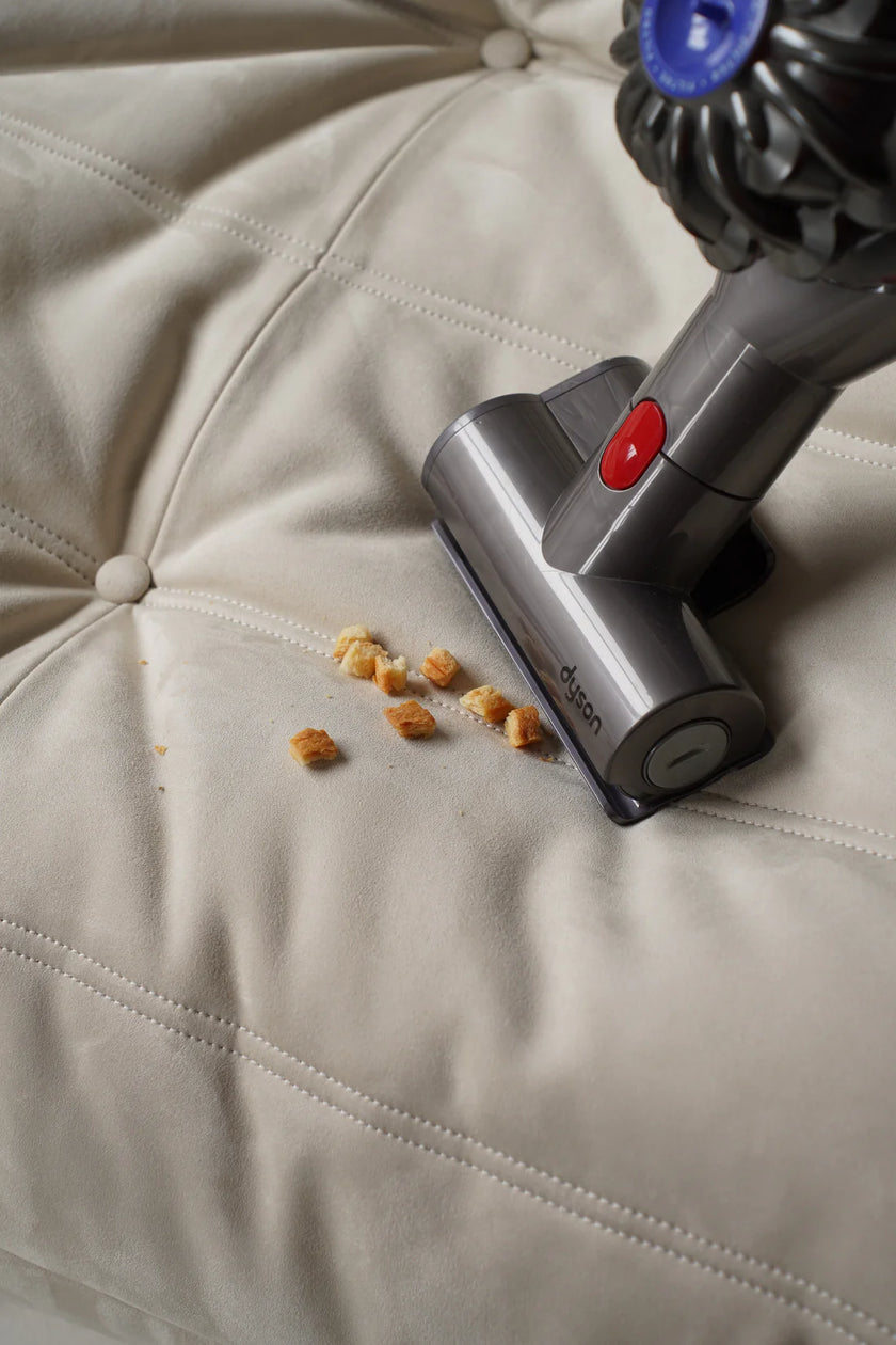 Dyson vacuum cleaner cleaning crumbs from a beige love seat sofa.