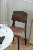 Wooden dining room chair with black legs near a round table with a glass and book on a wooden floor.