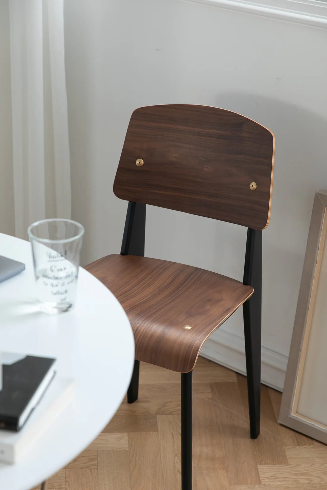 Wooden dining room chair with black legs near a round table with a glass and book on a wooden floor.
