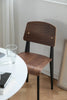 Wooden dining room chair with black legs near a round table with a glass and book on a wooden floor.