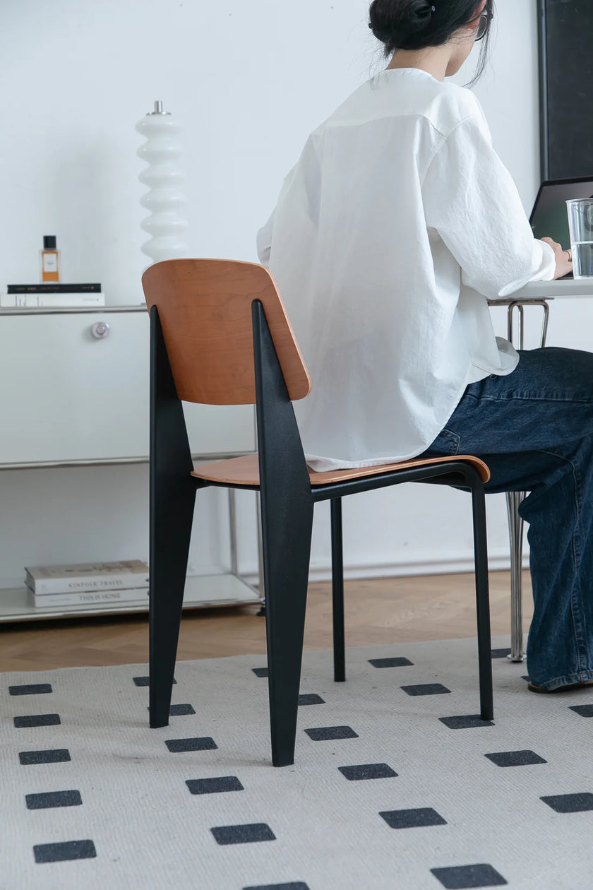 Lady sitting on a modern dining room chair with a brown seat and black legs in a room.