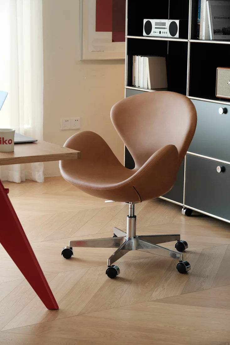 A modern swivel office chair with a curved, shell-like backrest upholstered in warm tan leather, mounted on a chrome five-star base with black casters. The chair is positioned in a bright, contemporary home office setting with light wood floors, a black and white storage cabinet, and a red table leg visible in the foreground.