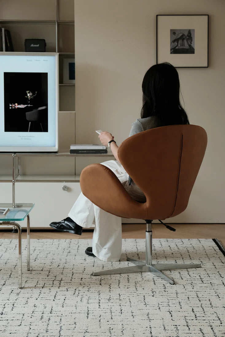 A woman sits in a brown egg-shaped swivel chair, viewed from behind, in a modern living room setting. She is wearing white pants and black shoes and holding a phone.
