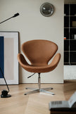 A classic mid-century modern Swan chair upholstered in warm tan leather with a chrome star base, set in a minimalist room with light wood floors, a black floor lamp, and framed wall art.