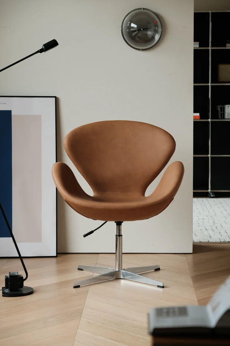 A classic mid-century modern Swan chair upholstered in warm tan leather with a chrome star base, set in a minimalist room with light wood floors, a black floor lamp, and framed wall art.