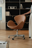 A modern tan leather swivel chair with a chrome five-star base and black casters, shown in a styled home office setting with a person touching the backrest.