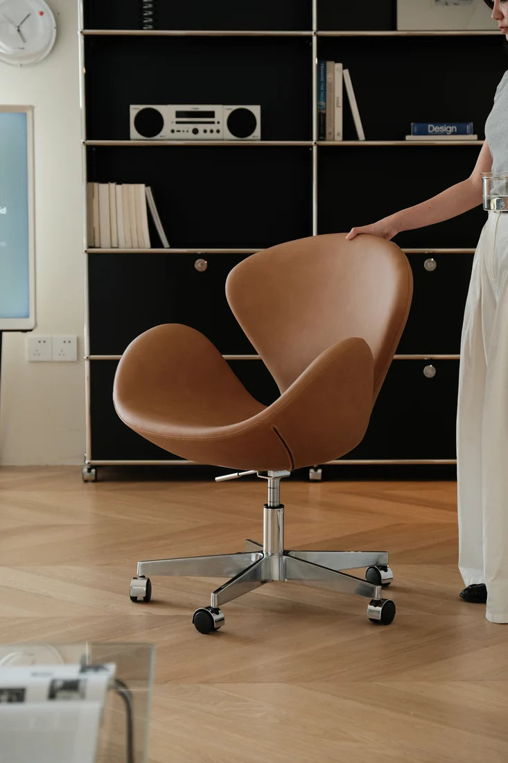 A modern tan leather swivel chair with a chrome five-star base and black casters, shown in a styled home office setting with a person touching the backrest.