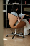 A woman sits in a tan leather swivel office chair with a chrome five-star base, reading a book in a modern room with wood floors and shelving.