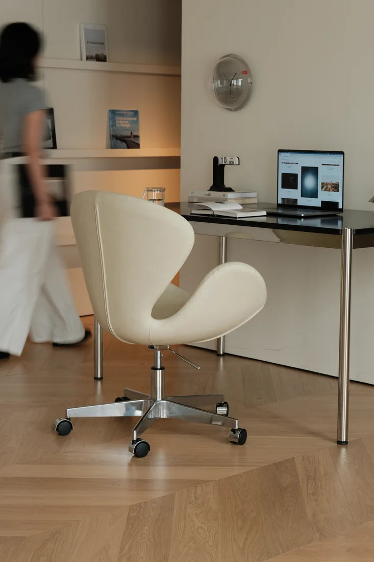 A modern cream-colored swivel office chair with a chrome five-star base and black casters, positioned in front of a black desk with a laptop in a minimalist home office setting.