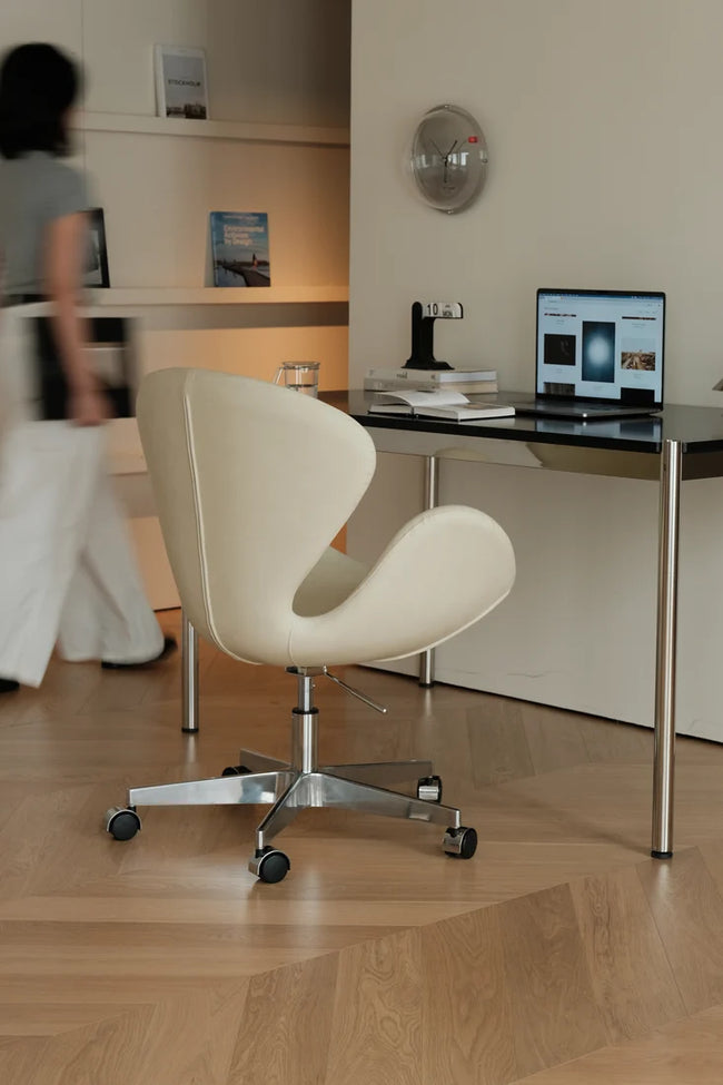 A modern cream-colored swivel office chair with a chrome five-star base and black casters, positioned in front of a black desk with a laptop in a minimalist home office setting.