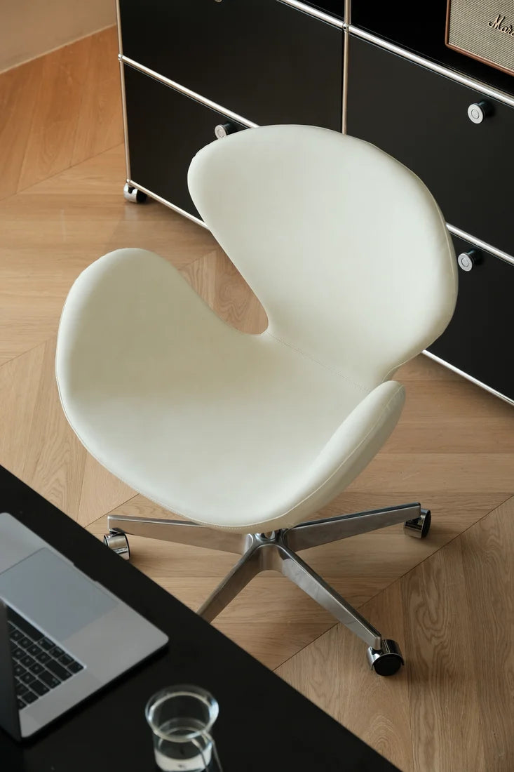 A modern white swivel office chair with a curved organic shell design and a chrome five-star base, positioned on a light wood herringbone floor next to a black desk.