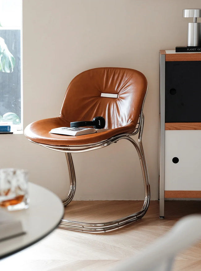 A mid-century modern accent chair with a curved brown leather seat and a polished chrome tubular metal base, styled in a minimalist room next to a cabinet.