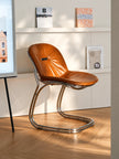A modern cantilever chair with a brown leather seat and a curved polished chrome base, set in a brightly lit room with light wood flooring and white wall shelves.
