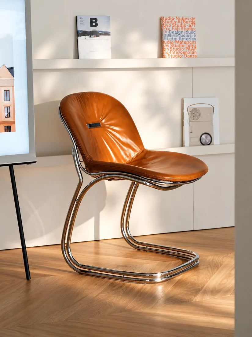 A modern cantilever chair with a brown leather seat and a curved polished chrome base, set in a brightly lit room with light wood flooring and white wall shelves.