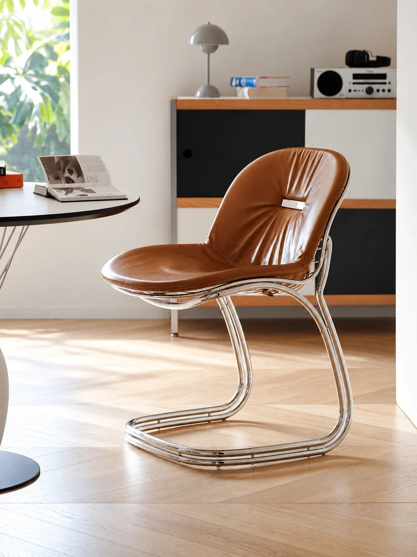 A modern side chair with a curved brown leather seat and a polished chrome tubular steel frame, shown in a bright, contemporary room setting.