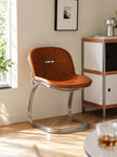 A mid-century modern accent chair with a brown leather seat and a curved chrome steel base, positioned in a brightly lit room next to a white and wood cabinet.