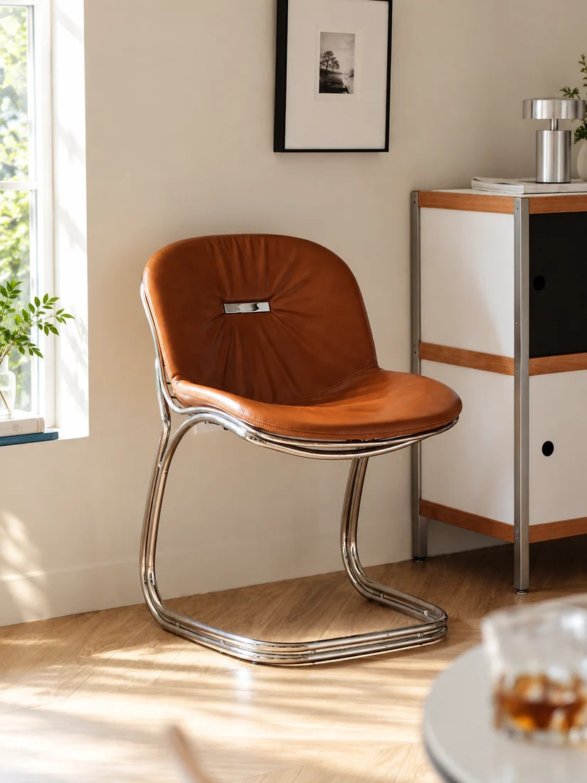 A mid-century modern accent chair with a brown leather seat and a curved chrome steel base, positioned in a brightly lit room next to a white and wood cabinet.