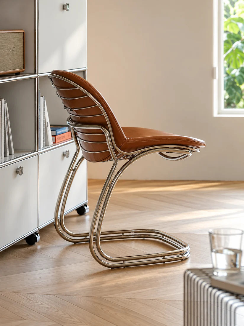 A modern cantilever chair with a brown leather seat and a curved polished chrome base, set in a brightly lit room with light wood flooring and white wall shelves.