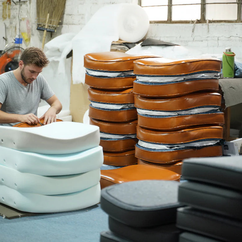 Man working on a leather seat in a workshop with stacks of leather seats and tools around.