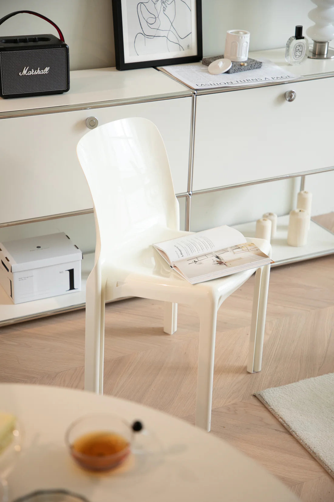 White living room chair with a magazine on a light wooden floor in a room with a desk and framed picture.