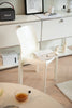White living room chair with a magazine on a light wooden floor in a room with a desk and framed picture.