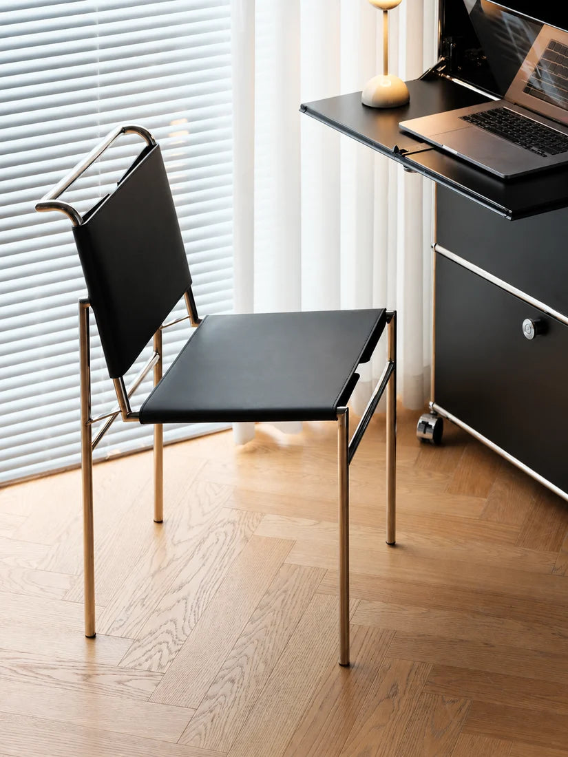 A modern black chair with a gold metal frame, positioned in a minimalist home office setting next to a black desk and laptop on a light wood herringbone floor.