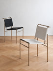 Two modern metal chairs with woven sling backs and seats, shown on a light wood herringbone floor against a white wall. One chair has a black seat and back, and the other has a white seat and back, both with gold-toned frames.